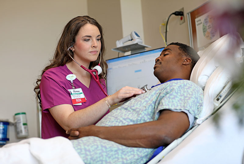 A nurse listens to a patient's heartbeat while they rest in a hospital bed.