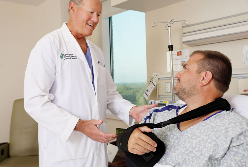 A smiling orthopaedics doctor speaking with a patient who is wearing an arm brace. 