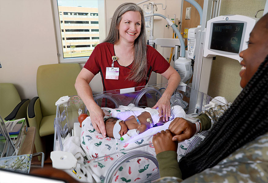 A nurse smiling at a mother. A newborn baby wearing an eye mask sleeps in a neonatal incubator.